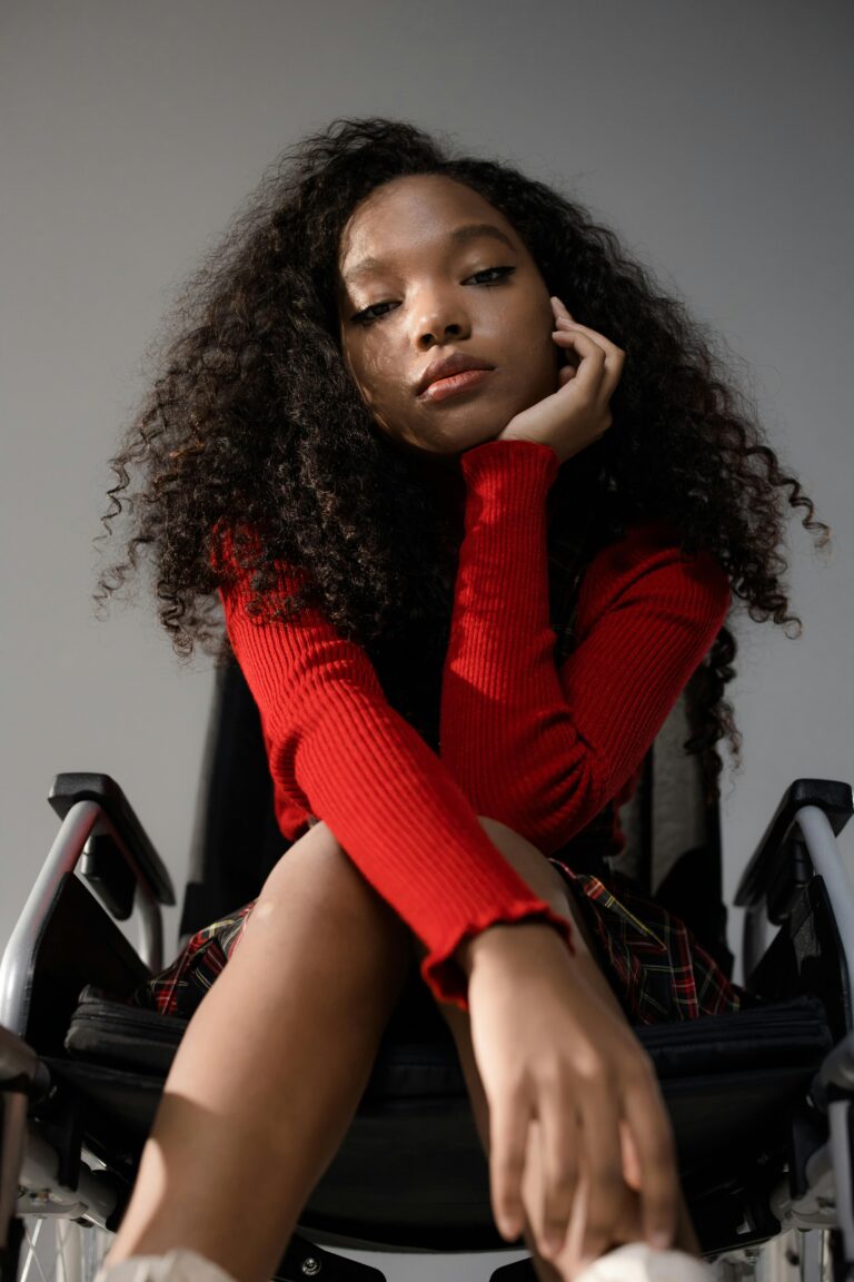 Portrait of a young woman with curly hair sitting in a wheelchair, wearing a red sweater.