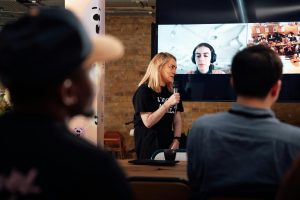 A woman presents to an audience during a hybrid conference in London, with virtual participants on screen.