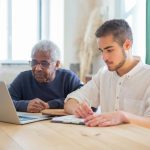 A senior man and young adult collaborating on a laptop at a table indoors, emphasizing technology use.